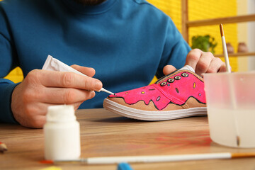 Man painting on sneaker at wooden table indoors, closeup. Customized shoes