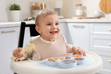 Cute little baby eating food in high chair at kitchen