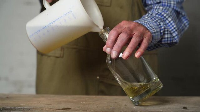 Older male hands are filling destilled yellow liquor from a plastic vessel into an empty glass bottle