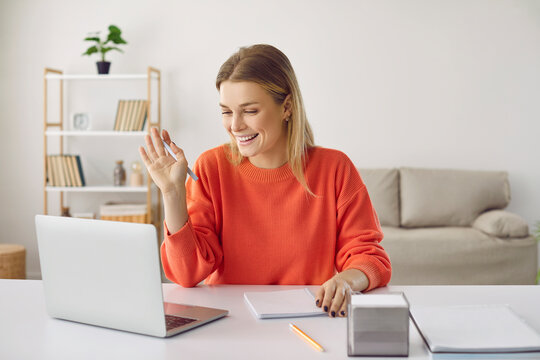 Happy student using laptop computer at working desk at home, having video conference, looking at screen, waving hand, talking to online business course teacher, learning new information, taking notes