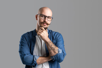 Portrait of thoughtful bald beard man in glasses who holds fist on chin isolated on gray background