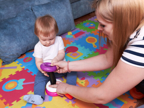 Mother with her child daughter play together seated on colorful playmat. Mom shows the child how to play correctly. Mom teaches her daughter to play educational toys.