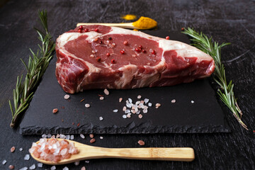 side view raw rib eye steak on a square black slate board with two spoons and rosemary. Minimalism.