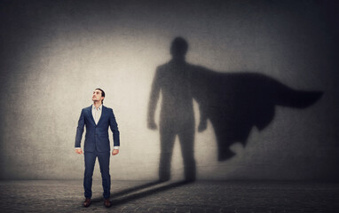Determined businessman stands confident in a hero stance and casting a brave superhero shadow on the wall behind. Business leadership and motivation concept. Ambition and strength symbols - obrazy, fototapety, plakaty