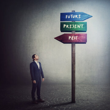 Businessman Stands In Front Of A Signpost Showing Three Different Directions, Past, Present And Future. Choose The Correct Way. Time Travel Road Sign. Hard Choice And Difficult Decision Concept