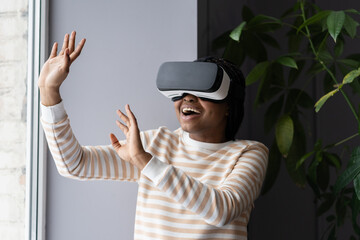 Overjoyed black woman in casual clothes enjoying spending leisure time in virtual reality while relaxing at home, happy black female sitting by window and playing VR games. Innovative technologies