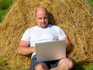 Naklejka na ściany i meble a middle-aged man, dressed in a T-shirt and shorts, is lying on a yellow haystack with a gray laptop in his hands. Remote work, training. Rural area.