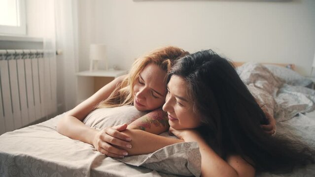 Homosexual lesbian couple lying in bed together. Take hands and smile each other with love. Family, woman love and romantic relationship. Beautiful asian girlfriend in morning home. LGBT pride event.