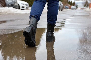 Female legs in black leather boots with reflection in puddle on a street. Rain in city, melting snow in early spring, waterproof footwear