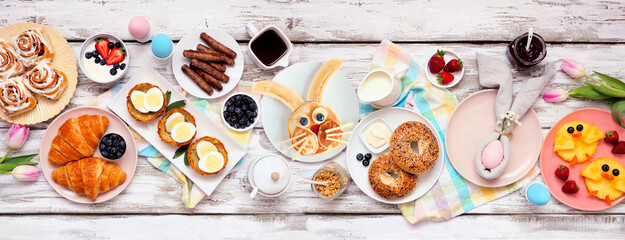 Easter breakfast or brunch table scene. Overhead view on a white wood banner background. Bunny pancake, egg nests, chick fruit and an assortment of spring food items. Copy space.