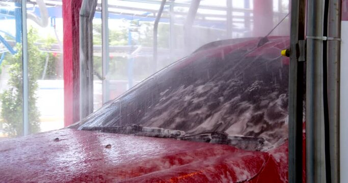 Red car moving though automatic car wash machine. Soapy water spray on the car