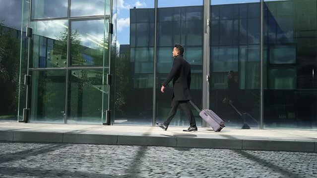 Young businessman holding suitcase and walking along glass building in modern city outdoors spbd. Side view of handsome guy moves luggage and walks confidently near office architecture, heads to