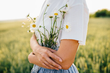 Gorgeous and creative caucasian woman decorate dress from chamomile cinching with belt around waste outfit, standing in wheat field at summer time in sunset. Woman unity nature concept.