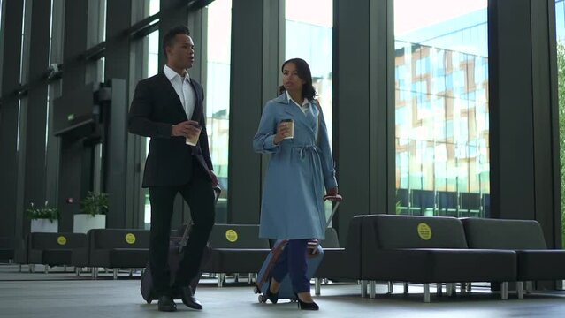 Young businesspeople or couple talking with coffee in hands and walking at airport spbd. African American businessman, businesswoman have dialogue and look with smiles, hold drinks and move luggage
