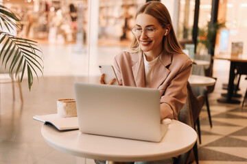 Portrait of a smiling student in headphones drinking coffee and working using a mobile phone and laptop in a cafe. A young business woman freelancer with glasses works remotely online from coffee shop