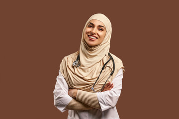Portrait of a female Muslim doctor wearing a white coat, arms crossed, standing against a brown...