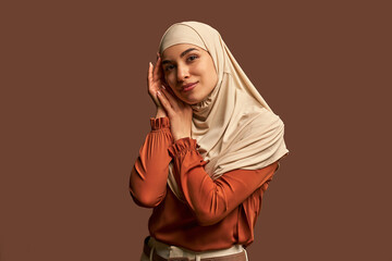 Modern, stylish, fashionable, beautiful Muslim woman dressed in a beige hijab, orange jacket and beige pants who looks at the camera and poses on a brown background.