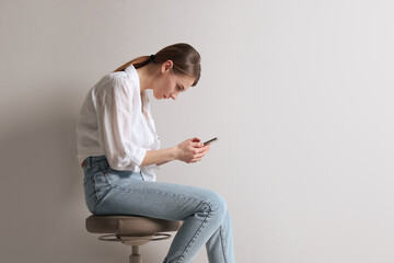 Woman with bad posture using smartphone while sitting on stool against light grey background, space...