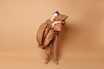 Sleepy woman with hair bun takes nap embraces pillow prepares for rest dressed in pajama sleepmask on eyes stands in full length against brown background. Good night and sleeping time concept
