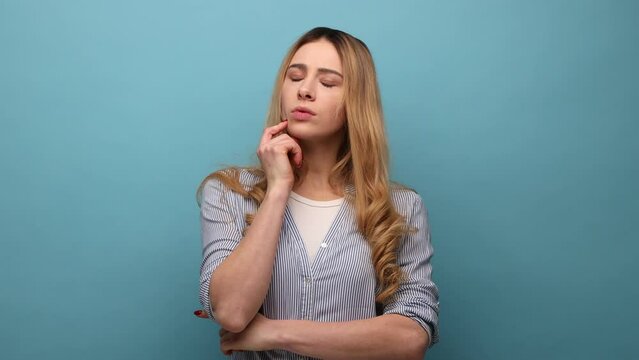 Portrait of pensive young adult woman with wavy hair thinking, holding chin, looking away, being deeply in thoughts, wearing striped shirt. Indoor studio shot isolated on blue background.