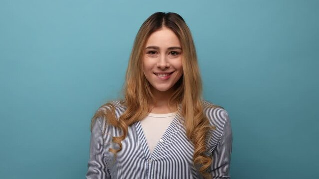 Portrait of positive optimistic blond woman standing and winking playfully, positive expression, looking at camera, wearing striped shirt. Indoor studio shot isolated on blue background.