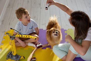 Mom, son and dog have fun playing a toy railroad and a toy crane in the nursery on puzzle mats. A...