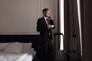 Handsome businessman drinking coffee while talking on phone near window in hotel room