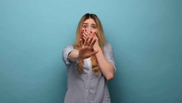 Portrait of scared blonde woman with wavy hair and raised arms, sees something awful, afraids, stop gesture, wearing striped shirt. Indoor studio shot isolated on blue background.