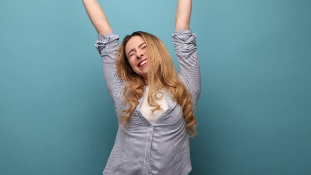 Portrait of victorious delighted happy woman raising hands, shouting for joy, screams celebrating win success, thrilled emotions, wearing striped shirt. Indoor studio shot isolated on blue background.