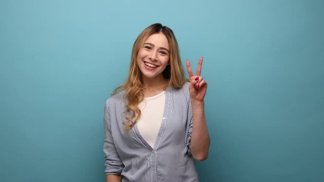 Portrait of happy attractive excited woman smiling and gesturing victory sign at camera, success or achievement, wearing striped shirt. Indoor studio shot isolated on blue background.