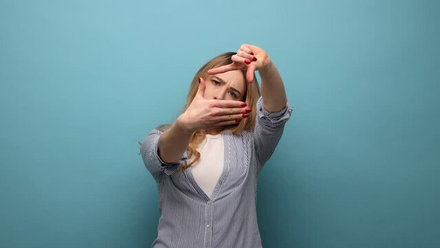 Portrait of blond woman imitating to take picture, looking through finger frame with attentive view, focusing and cropping image, wearing striped shirt. Indoor studio shot isolated on blue background.