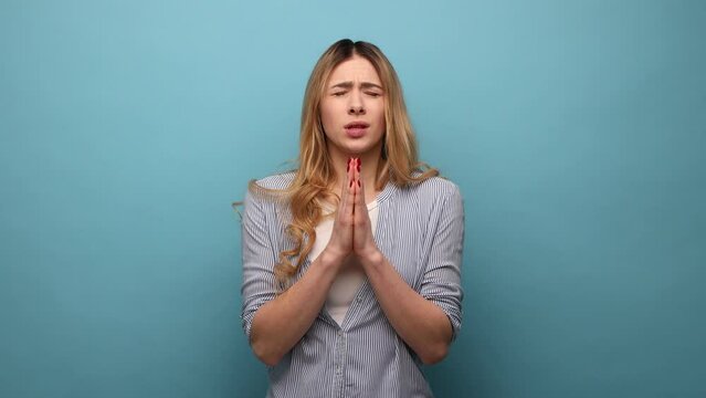 Woman with wavy hair holding hands in prayer and pleading, looking up with eyes full of hope, asking apology, wearing striped shirt. Indoor studio shot isolated on blue background.