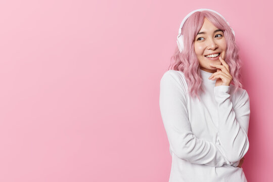 Happy thoughtful Asian woman smiles pleasantly listens audiotrack via stereo headphones wears casual white jumper isolated over pink background with copy space away. Listening favorite tunes