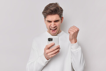 Outraged man looks angrily at smartphone gets annoying message clenches fist and exclaims loudly wears casual jumper isolated over white background argues with phone expresses negative emotions