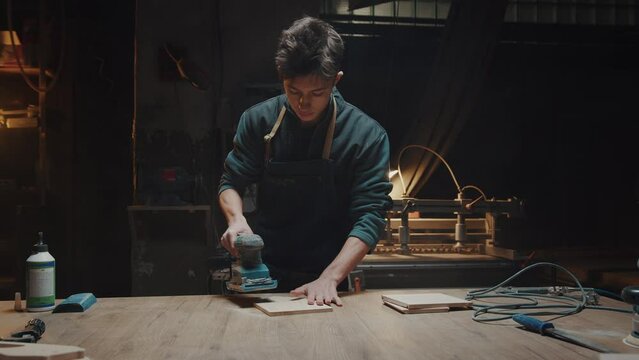 Young man works in his workshop, polishing wooden parts and blanks. Carpenter and his small family business. Handmade wooden products