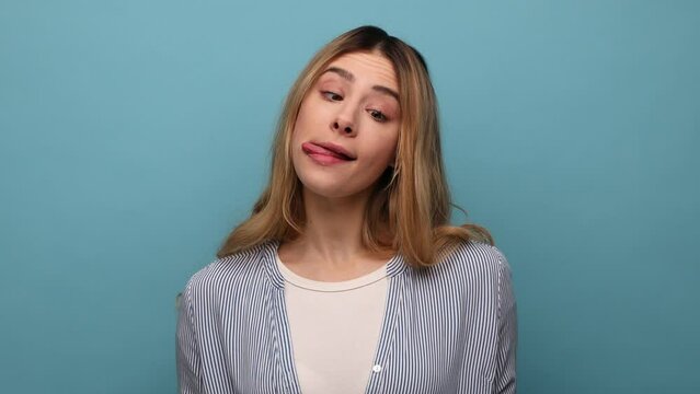 Funny young woman crossing eyes and showing tongue out, playing fool, making silly balmy face, brainless dumb expression, wearing striped shirt. Indoor studio shot isolated on blue background.