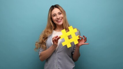 Smiling beautiful woman holding yellow hashtag symbol, making important topic popular, setting trends on Internet, wearing striped shirt. Indoor studio shot isolated on blue background.