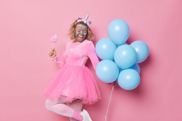 Obraz na płótnie Canvas Overjoyed woman applies clay mask on face for rejuvenation dances carefree and sings has good mood during holiday wears unicorn hedband and dress holds bunch of blue helium balloons. Celebration
