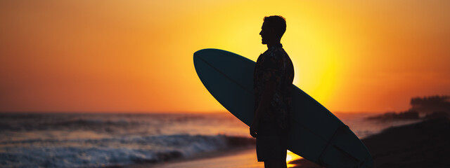 Silhouette of surfer in hawaiian t-shirt with surf board standing on the beach at sunset and looking far, panoramic - obrazy, fototapety, plakaty