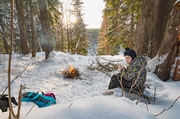 A boy sitting by a campfire in the woods in winter. - 488421623