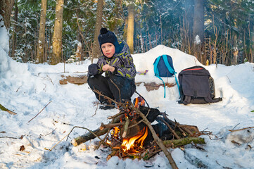 A boy sitting by a campfire in the woods in winter. - 488421624