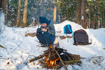 A boy sitting by a campfire in the woods in winter. - 488421625