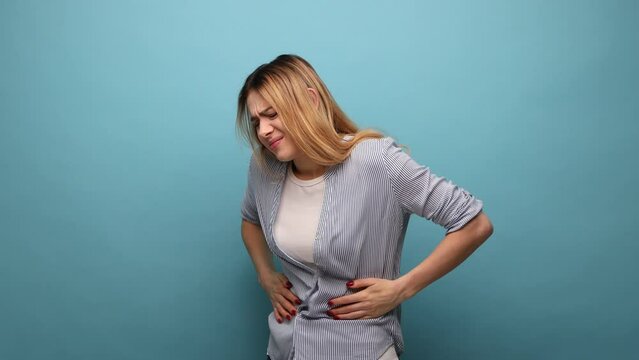 Portrait of unhealthy distressed woman clutching her belly, grimacing from acute abdominal pain, gastritis or constipation, wearing striped shirt. Indoor studio shot isolated on blue background.