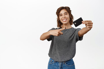 Fototapeta premium Portrait of beautiful young woman showing credit card, recommending bank, contactless shopping or discounts in store, time to shop, standing over white background