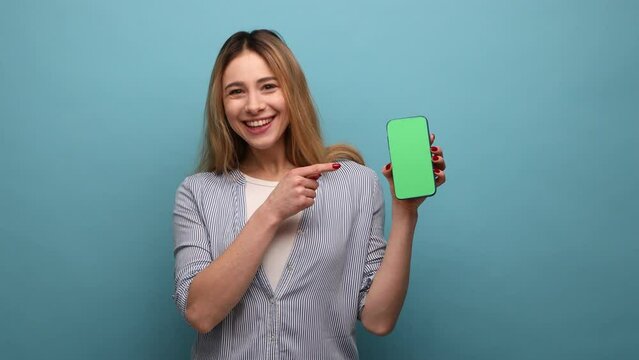 Look at my cellphone. Adorable glad woman pointing smartphone with finger and smiling at camera, advertising of mobile device, wearing striped shirt. Indoor studio shot isolated on blue background.