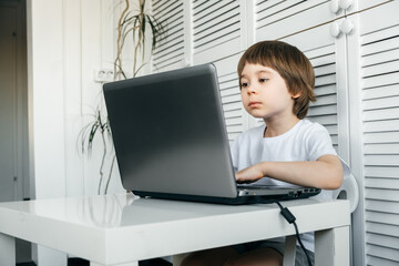 5 year boy sit at the table, uses laptop, looking at the screen. Child is doing homework lesson, playing video games, studying IT development, watching cartoon. Home distance online education concept