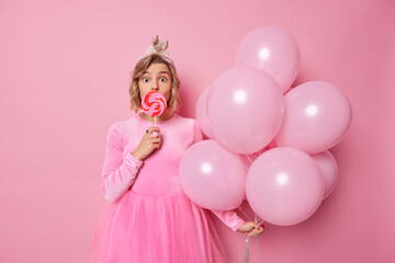 Obraz na płótnie Canvas Surprised young woman looks wondered at camera covers mouth with heart shaped caramel candy wears festive dress holds bunch of helium balloons isolated over pink background. Festive occasion