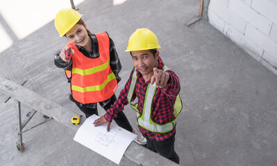 Fototapeta premium Top view of two Asian civil engineers wearing safety hardhat standing looking blueprint and pointing while checking infrastructure in the construction site. 