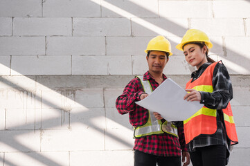 Two Asian civil engineers wear safety hard hats standing hold and look blueprint while checking...