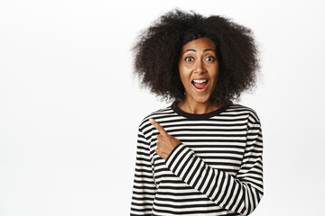 Close up of enthusiastic african american woman pointing finger left, smiling and showing advertisement, announce promo sale, demonstrate information aside, white background